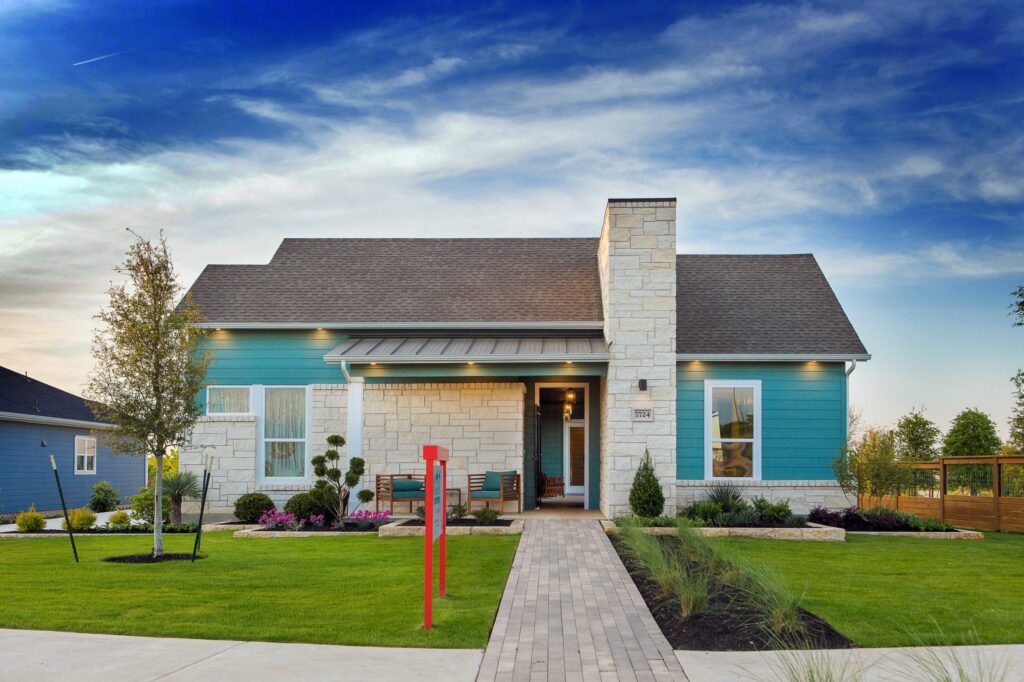 A modern single-story home with teal siding, stone accents, a covered front porch, landscaped yard, and a tall stone chimney under a dramatic evening sky.